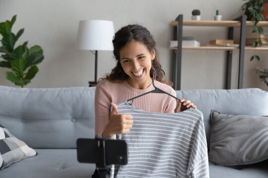 Smiling Caucasian girl influencer vlogger record clothes unpacking on smartphone camera at home, happy young woman stylist blogger recommend apparel, satisfied with online shopping in internet