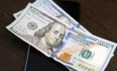 One hundred dollar bills and smartphone lie on a wooden table. Mobile banking concept. Close-up.