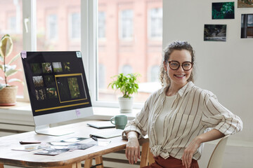 Portrait of young female photographer smiling at camera while posing at desk with photo editing...