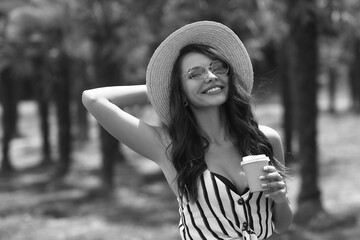 Closeup breast up portrait of young beautiful smiling girl in shirt, salty hat and sunglasses in palms. Happy joyous female model. Tanned girl drinking coffee