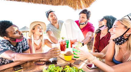 Young multiracial friends drinking at beach cocktail pub with open face mask - New normal summer concept with milenials having fun together toasting drinks - Bright filter with focus on central people