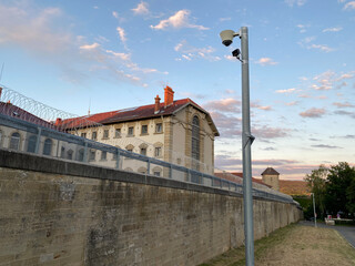 Gefängnismauer mit Stacheldrahtringe auf der Spitze, Dahinter pompöses historisches Gefängnisgebäude mit rotem Dach im Morgenlicht
