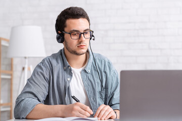 Webinars and trainings. Focused guy with glasses and headphones makes notes in notebook and looks at laptop