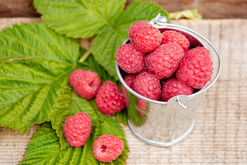 Raspberries in an iron bucket, green leaves and a scattering of berries on a wooden table. Top view. Harvest concept.