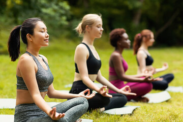 Concentrated multinational ladies meditating or doing breathing exercises on yoga practice outside,...