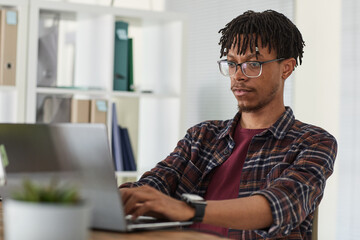 Portrait of contemporary African-American man using laptop and typing while working at desk in...