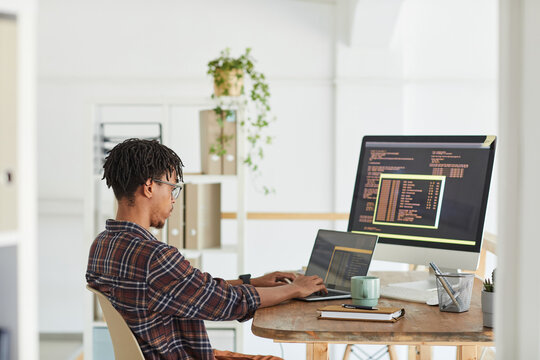 Side view at African-American IT developer typing on keyboard with black and orange programming code on computer screen and laptop, copy space