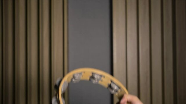 Caucasian adult male hand playing and shaking circular tambourine percussion in recording studio with wall in background, close up static