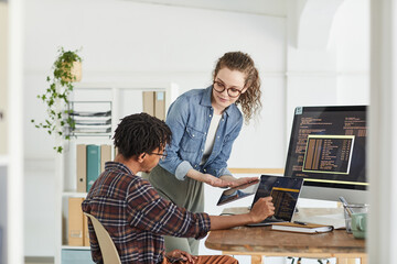 Portrait of smiling young woman talking to African-American man writing code while working in web...