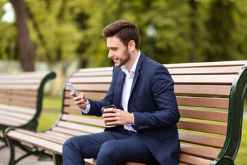 Obraz premium Handsome office employee having coffee to go and browsing web via smartphone on bench at park