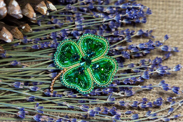 Handmade beaded brooch in the form of a four-leaf clover against a background of dry lavender branches, autumn leaves. A symbol of good luck and wealth in Irish mythology.