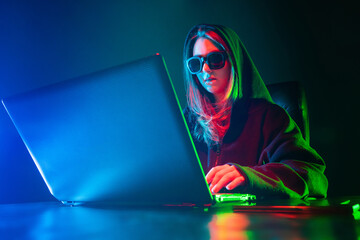 Teenagers hobbies. A teenager watches a video on the Internet. A girl with dark glasses is sitting at a laptop. The girl at the computer is illuminated by colored neon lights.