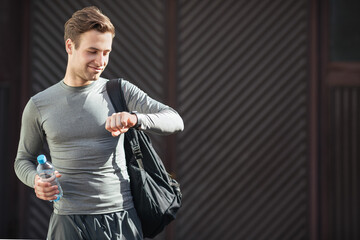 Young smiling man goes to training with sports bag and in sportswear, looks at tracker on hand