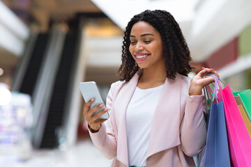 Discount App. Smiling Black Woman With Shopping Bags Using Smartphone In Mall