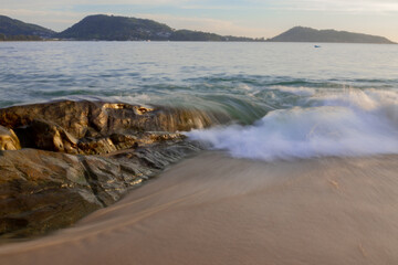 A view of a sandy beach with a large stone on the shore about which waves are beating, long exposure.