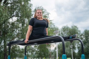 Obraz premium A beautiful smiling overweight young woman stretches for split on uneven bars outdoors. Fat girl gymnast doing fitness on the sports field. Sport outside on a warm summer day.
