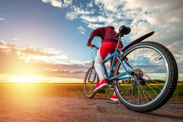 Fototapeta na wymiar Beautiful girl in a red sports suit on a bicycle on a sunset background. The concept of a healthy lifestyle, sports training, cardio load. Copy space.