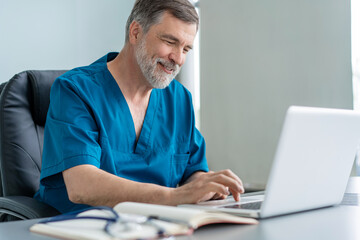 Mature male doctor working on laptop computer, sitting in medical office.