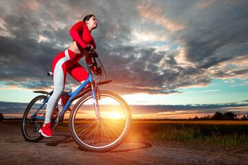 Naklejka na ściany i meble Beautiful girl in a red sports suit on a bicycle on a sunset background. The concept of a healthy lifestyle, sports training, cardio load. Copy space.