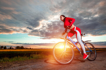 Fototapeta na wymiar Beautiful girl in a red sports suit on a bicycle on a sunset background. The concept of a healthy lifestyle, sports training, cardio load. Copy space.