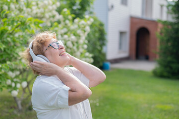 An elderly Caucasian woman walks in a park and listens to music. A smiling grandmother enjoys the aroma of flowering trees in the garden on a warm sunny day.