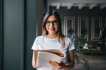 Young smiling caucasian woman in glasses standing near the window in business hall, taking notes in a notepad.
