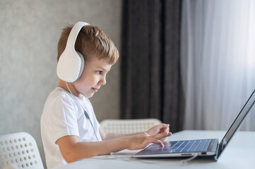 Cute diligent boy is typing on the keyboard and is carefully looking at the laptop screen. Caucasian schoolboy sits at a table, listens to music on headphones and does homework. Distance education.