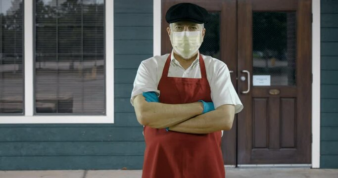 The Owner Of A Small Bakery Or Butcher Shop Stands In Front Of His Store Wearing A Mask Due To COVID19  Mandates.