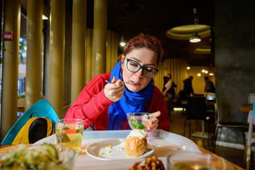 Red-haired caucasian woman eating salad in a cafe. Pensive romantic girl in glasses and a blue scarf is having lunch.