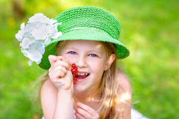 Close-up portrait of a 6-year-old blonde girl eating red currants. A girl in a green hat with a flower. 