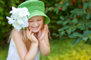 A little blond girl laughs with her eyes closed. A girl in a green hat with a big white flower