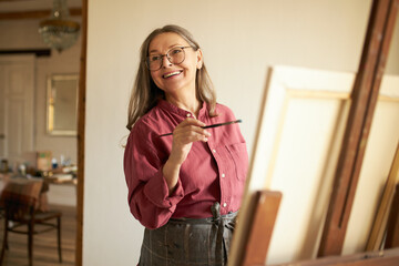 Fototapeta Attractive cheerful female pensioner in apron and eyeglasses imroving artistic skills, standing in studio next to easel, holding paintbrush during art workshop. People, leisure and hobby concept obraz
