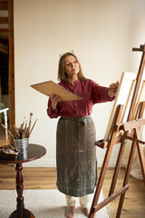 Full length vertical shot of inspired mature female painter working in her studio wearing apron, standing barefooted in front of easel, applying oil paint on canvas. Creativity and imagination