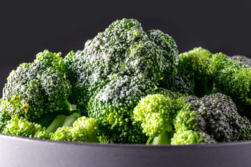 close up view of fresh and green broccoli in a bowl, side view on dark black background, spilled water on top of the vegetables