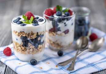 Granola with white yogurt with raspberries and blueberries in two glasses on blue napkin.