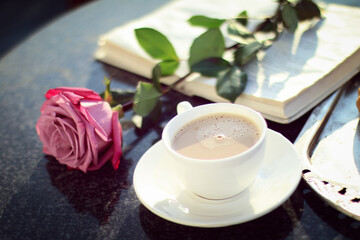 cup of cappuccino on a table next to an open book and a rose