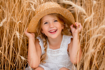 Cute little blonde girl in a straw hat playing in a wheat field on a warm summer day