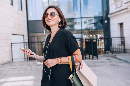 Smiling beautiful modern middle-aged female Portrait dressed black dress and sunglasses with shopping bags holding slim smartphone listening music with wired earphones. Beautiful people concept image.