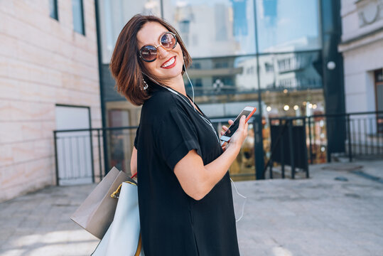 Beautiful modern middle-aged female Smiling Portrait dressed black dress and sunglasses with shopping bags holding slim smartphone listening music with wired earphones. Beautiful people concept image.
