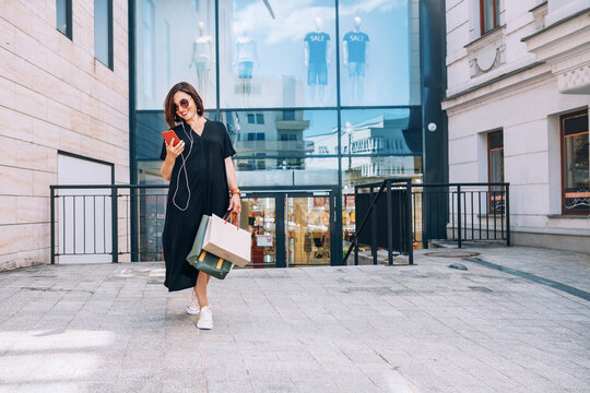 Smiling beautiful modern middle-aged female Portrait dressed black dress and sunglasses with shopping bags browsing slim smartphone listening music with wired earphones. Beautiful people concept image