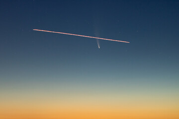 A plane flying in front of C/2020 F3, or Comet Neowise, in the early morning sky with a gradient from orange sun glow to blue night sky. 