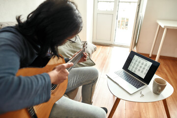 Learning guitar in self-isolation. Young man sitting on sofa at home and learning how to play...