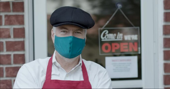 The owner of a small bakery or butcher shop wearing a mandatory mask due to COVID19 comes out and stands in front of his store.