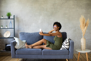 Obraz premium Happy african American young woman sit relax on cozy couch happy to move to new apartment. Smiling black girl rest on comfortable sofa in living room dreaming