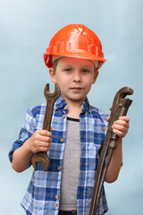 A preschooler boy is standing and holding a wrench and an adjustable wrench. on a blue background. helmet on the head. Concept - young builder, plumber, assistant, locksmith. Vertical photo.