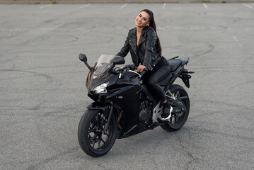Fototapeta na wymiar Attractive girl with long hair in black leather jacket and pants on outdoors parking with stylish sports motorcycle at sunset.