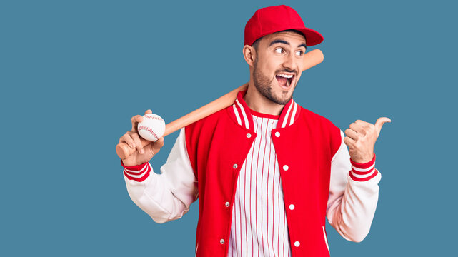 Young handsome man holding baseball bat and ball pointing thumb up to the side smiling happy with open mouth