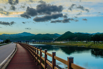Car traveling on two lane country road along lakeside boardwalk under blue sky with large white clouds at sunset.