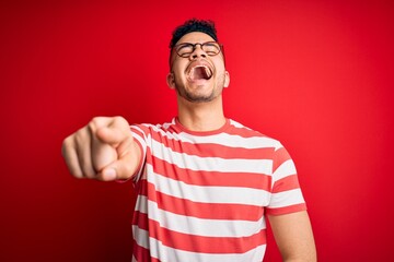 Naklejka premium Young handsome man wearing casual striped t-shirt and glasses over isolated red background laughing at you, pointing finger to the camera with hand over body, shame expression