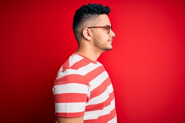 Young handsome man wearing casual striped t-shirt and glasses over isolated red background looking...